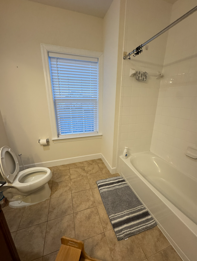 A bathroom featuring a toilet, a bathtub with a shower curtain rod, a window with blinds, and a striped rug on tiled floors.