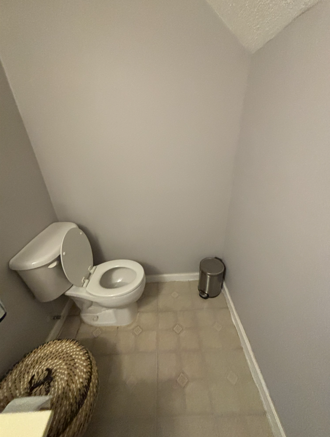 A light gray restroom featuring a white toilet, a small silver pedal trash can, and a woven basket in the foreground.