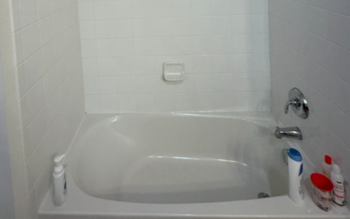A bathroom with a white toilet, tiled floor, a window with blinds, and a white bathtub with a shower curtain rod.