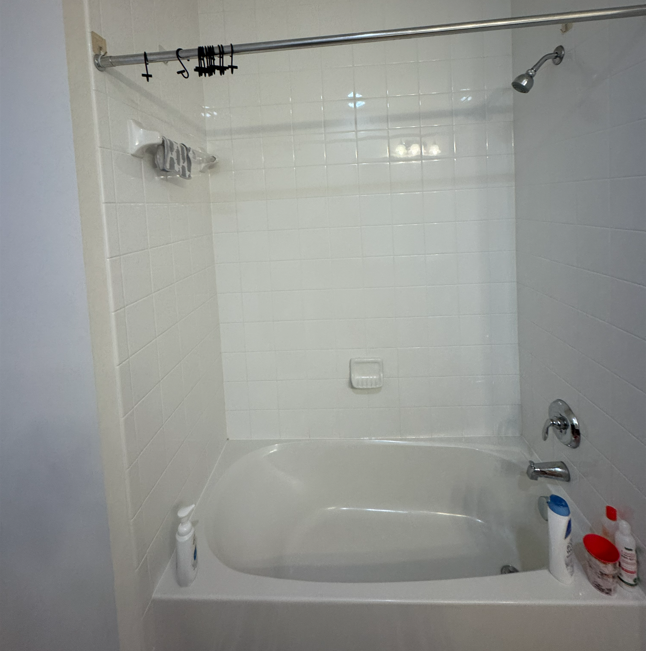 A white bathtub with a showerhead and a curtain rod with rings, against tiled walls, with toiletries on the ledge.