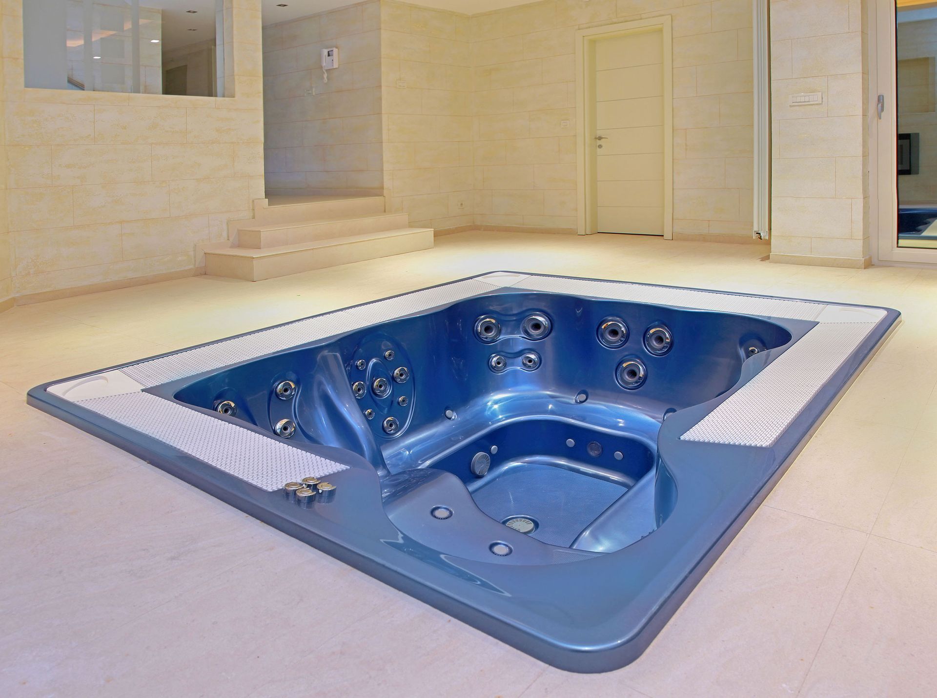 A rectangular blue hot tub is set into the floor of a spacious, light-colored room with tiled walls and steps.