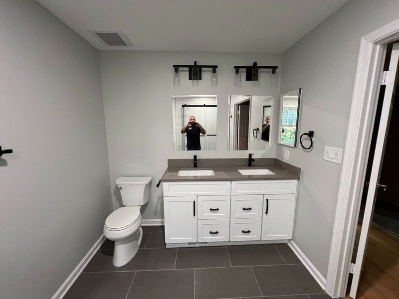Bathroom with a white vanity, grey walls, dark floor tiles, toilet, two mirrors, and dark fixtures.