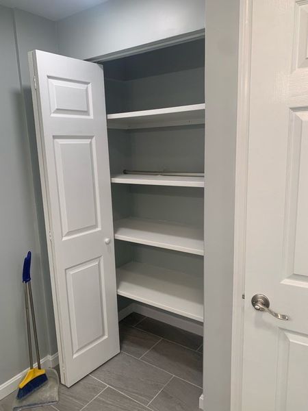 White closet with shelves and door ajar, broom in the corner, on gray tiled floor.