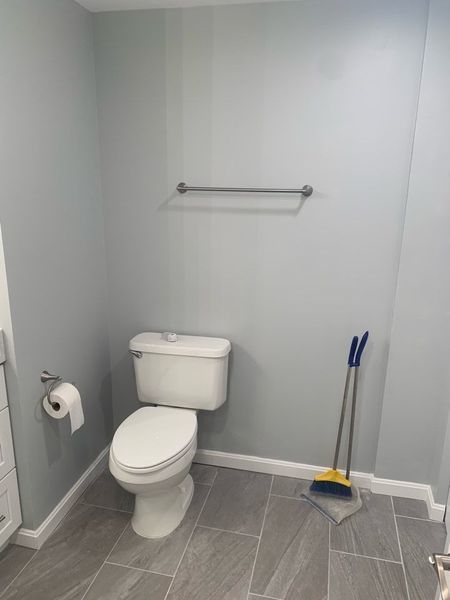 Bathroom with a toilet, towel rack, and broom and dustpan. Gray walls and flooring.