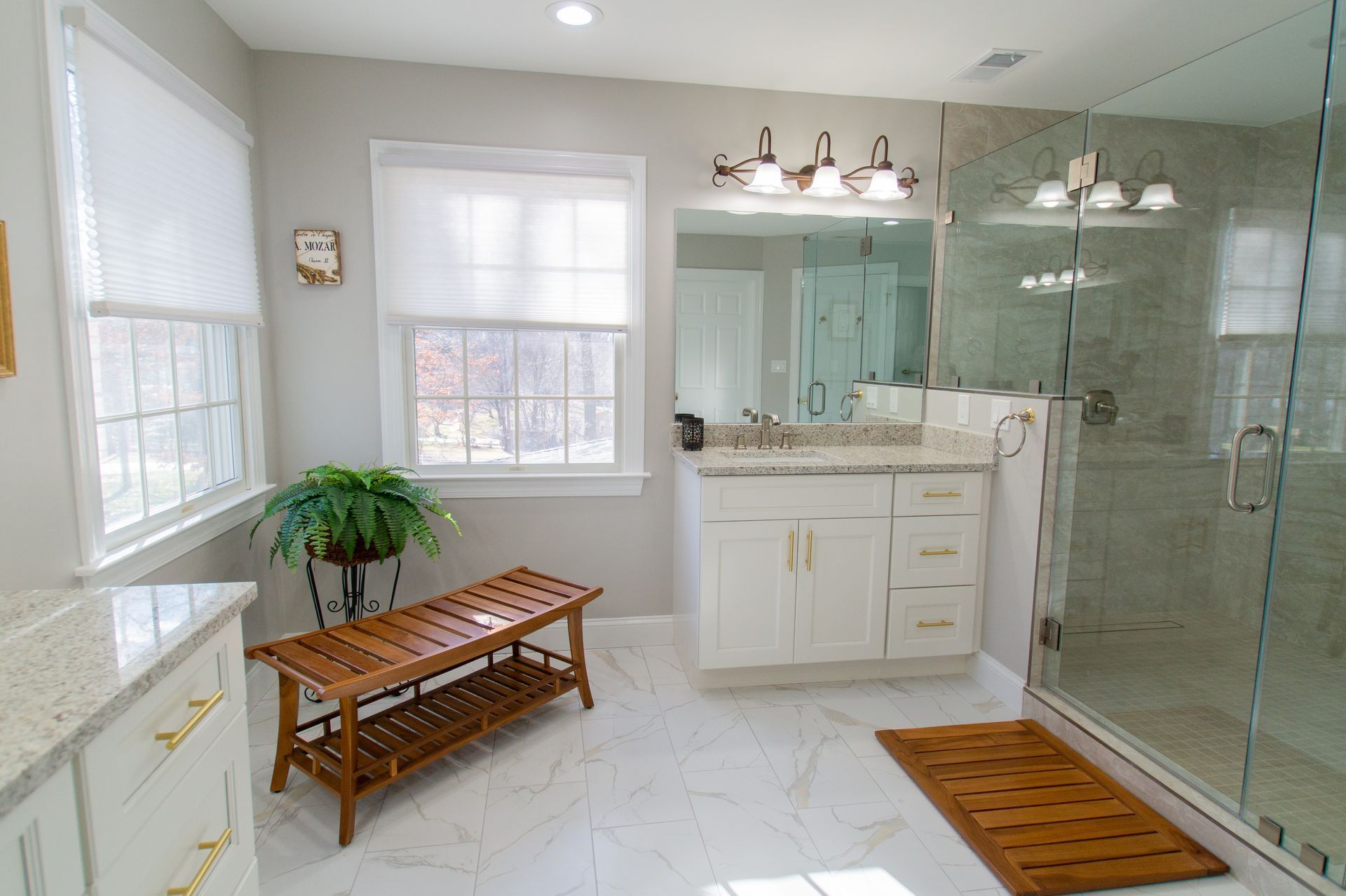 Bright bathroom with white walls, cabinets, and a glass shower. Includes a wooden bench, rug, and green plant.