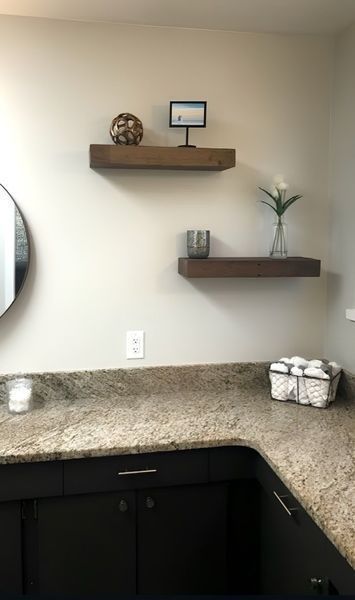 Bathroom with floating shelves. Shelves hold decorative items. Beige walls, dark cabinets, and granite countertop.
