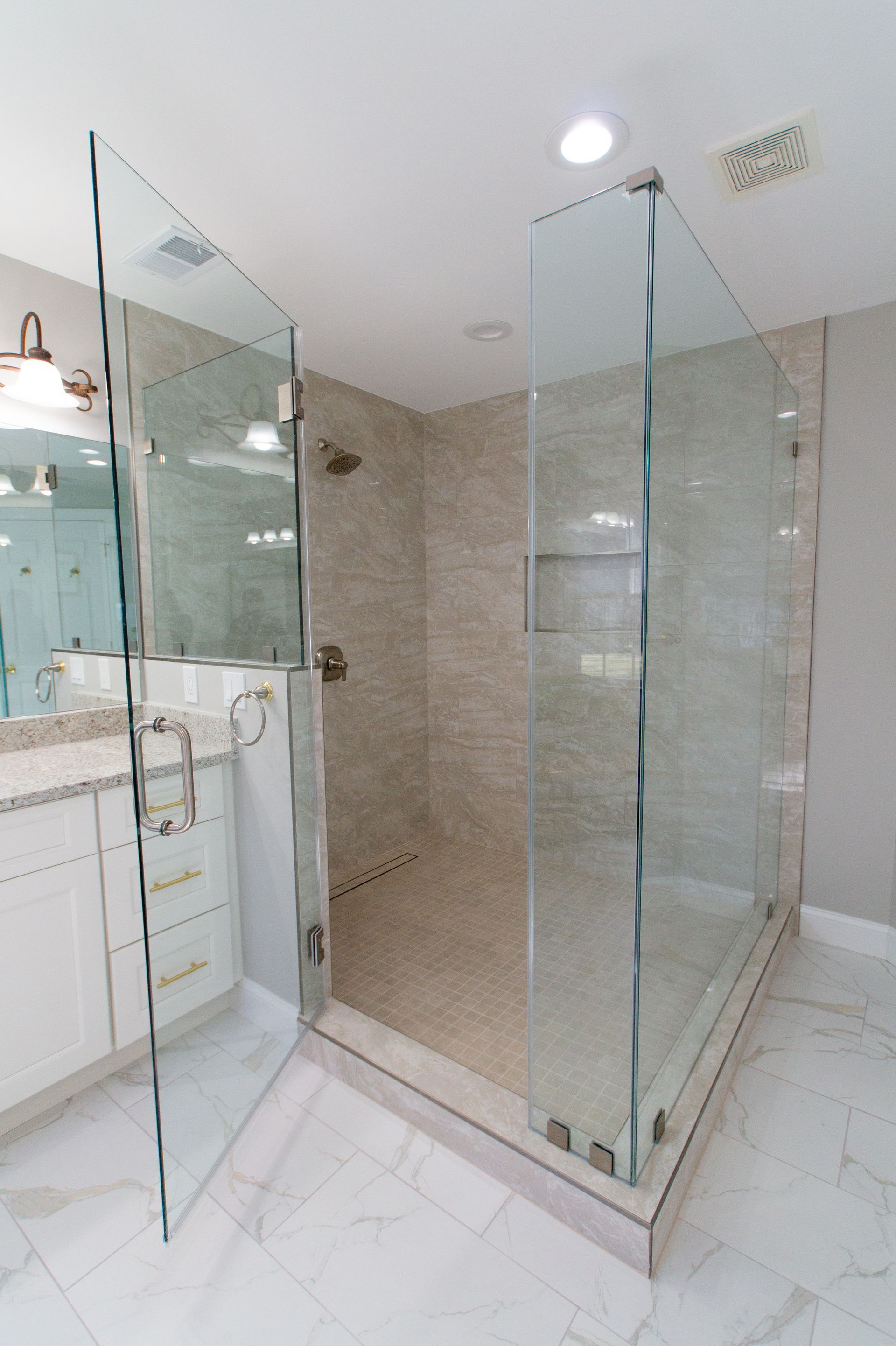 Glass-walled shower with light-colored tile walls and floor, and an open door next to a vanity in a bathroom.