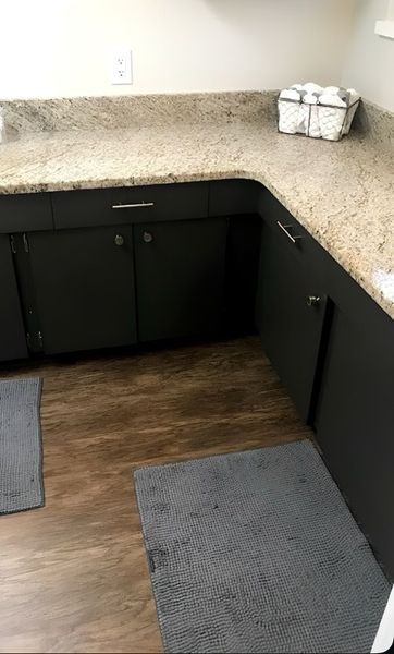 Dark cabinets with silver hardware below a tan countertop in a kitchen; gray rugs on a wood floor.