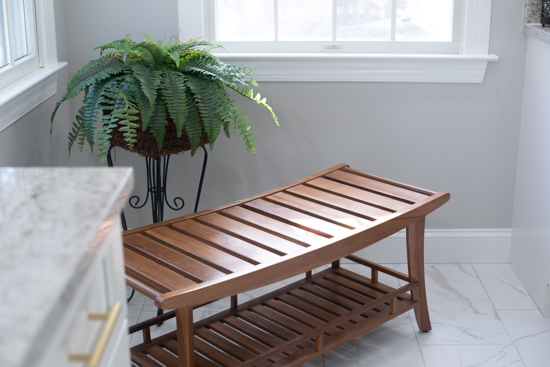 Wooden bench and fern plant near a window in a bathroom with light gray walls and white tile floor.