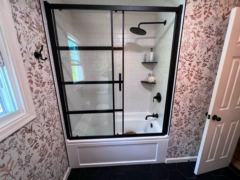 Bathroom with black-framed shower, white tub, and patterned wallpaper. Door propped open.