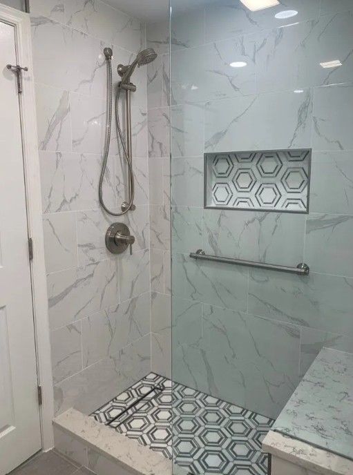 Shower with marble-patterned tile and a glass door. Built-in shelf has patterned tile; a grab bar is installed.