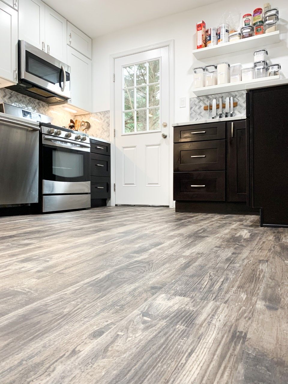 Kitchen with wood-look flooring, stainless steel appliances, dark cabinets, and a white door leading outside.