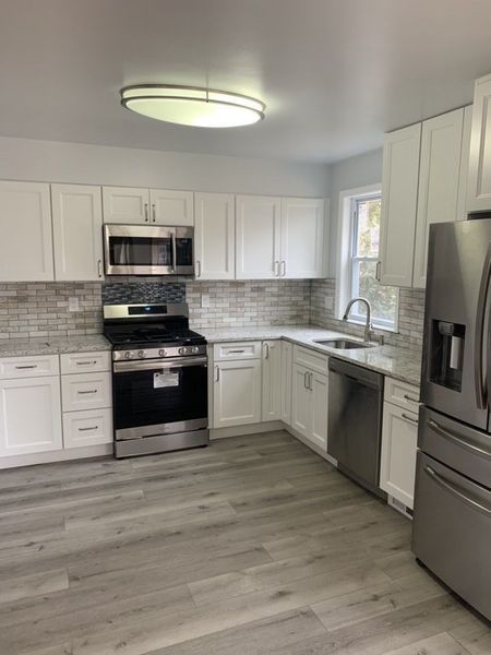 White kitchen with stainless steel appliances, grey flooring, and a light-colored brick backsplash.