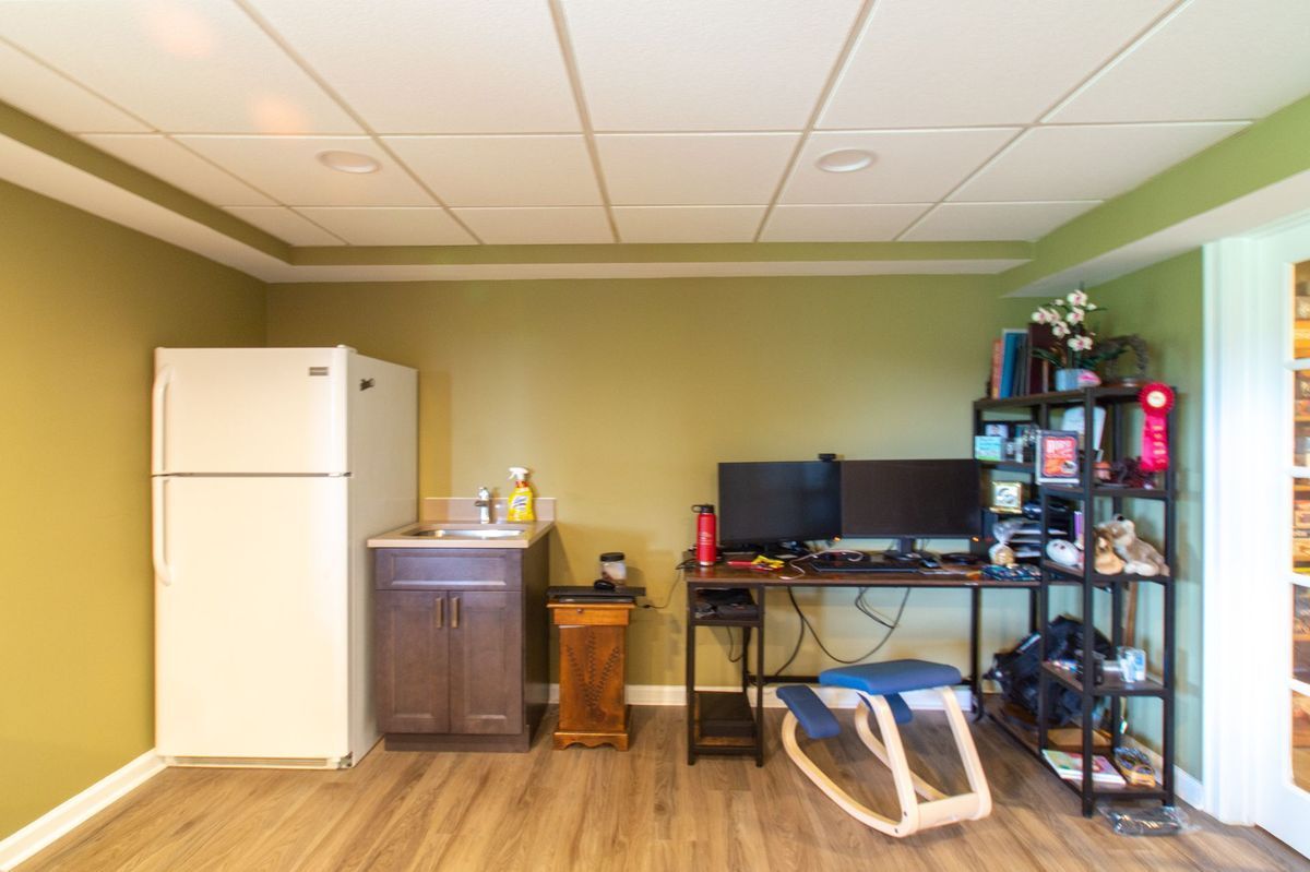 Room with desk, fridge, and shelving unit against olive green walls and wood-look flooring.