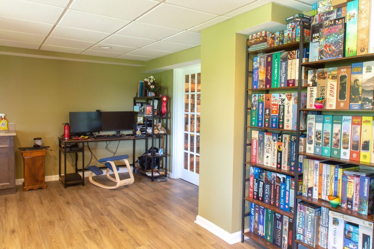 Game room with board game shelves, desk with dual monitors, and wooden floors.