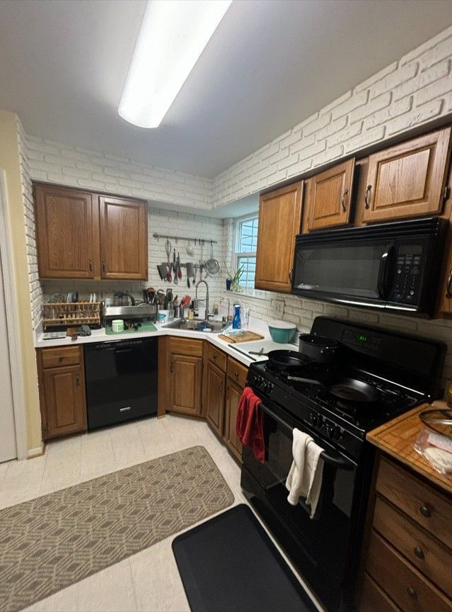 A kitchen with white brick walls, wood cabinets, a black stove, a black dishwasher, and two floor mats on a light floor.