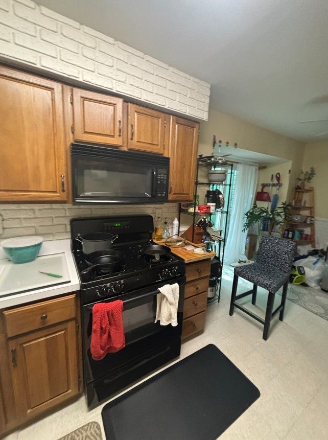 A kitchen with wood cabinets, a black stove and microwave, a white brick backsplash, and a tall stool in the background.