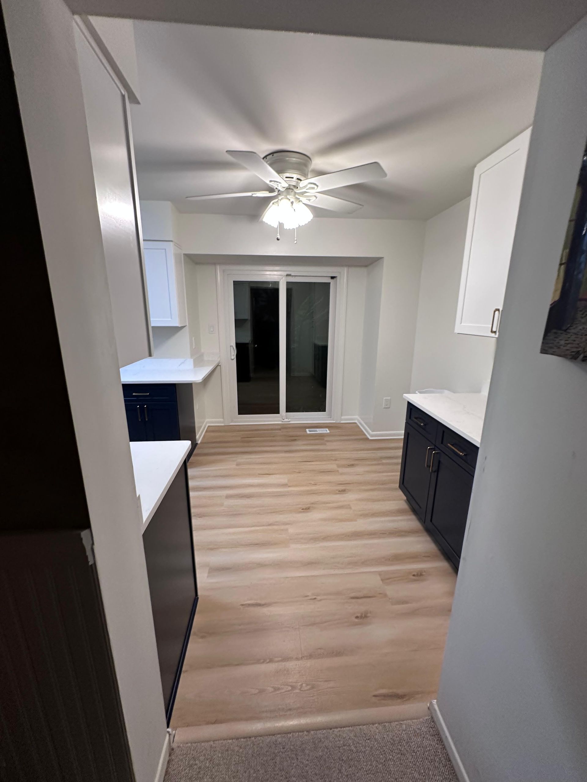 A kitchen with white upper cabinets, dark blue lower cabinets, light wood floors, and a ceiling fan over a sliding door.