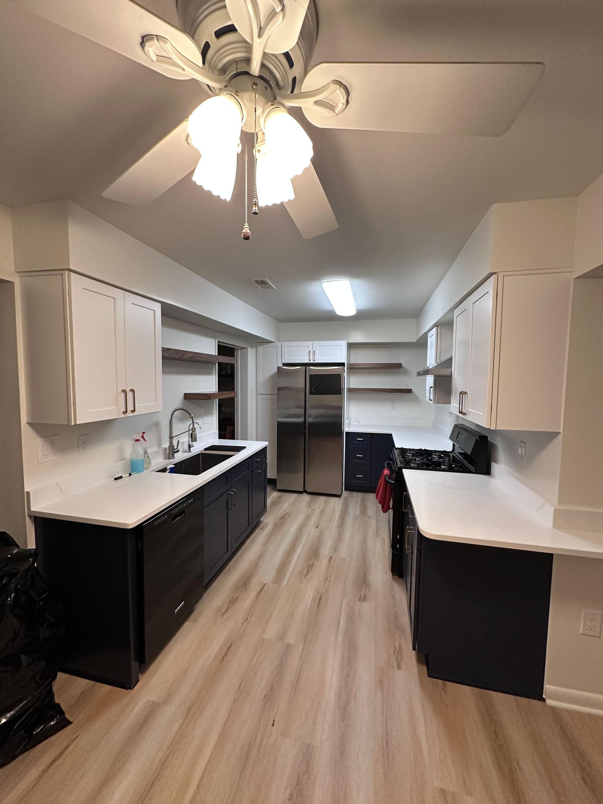 A galley-style kitchen with white upper cabinets, dark base cabinets, white countertops, stainless steel appliances, and wood floors.