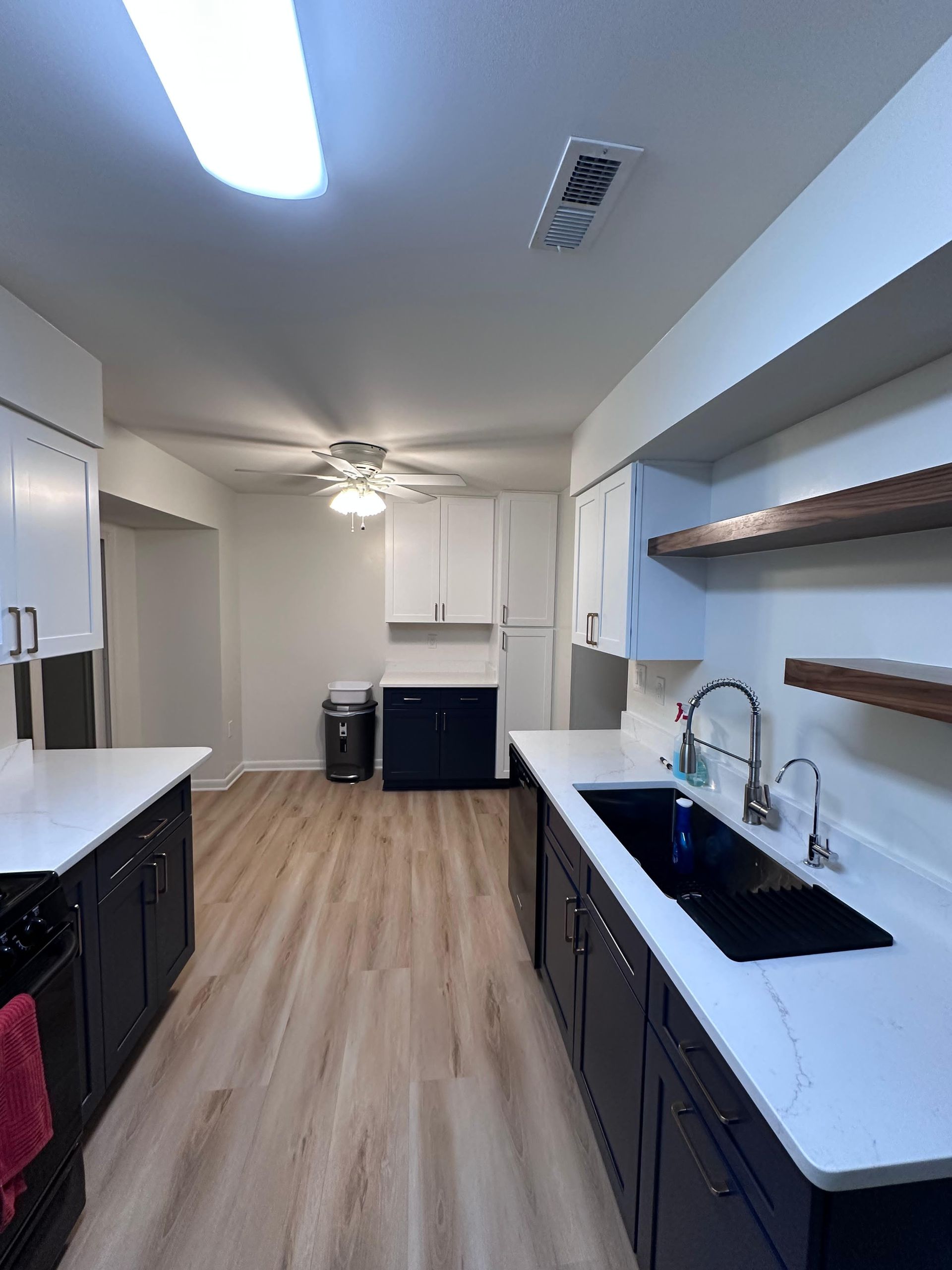 A galley kitchen with dark lower cabinets, white upper cabinets, light wood-look flooring, and white countertops.