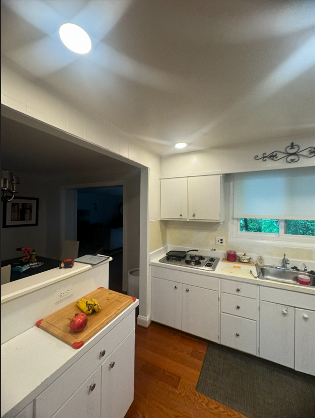 A bright kitchen with white cabinets, hardwood floors, a stove, a sink, and a wooden cutting board with fruit on a counter.