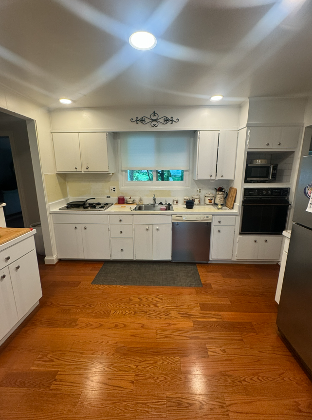 A brightly lit kitchen with white cabinets, stainless steel appliances, and wood floors featuring a grey rug by the sink.