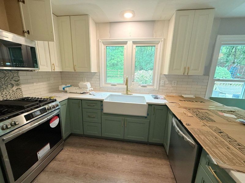 Kitchen with sage green cabinets, white backsplash, farmhouse sink, and white upper cabinets.
