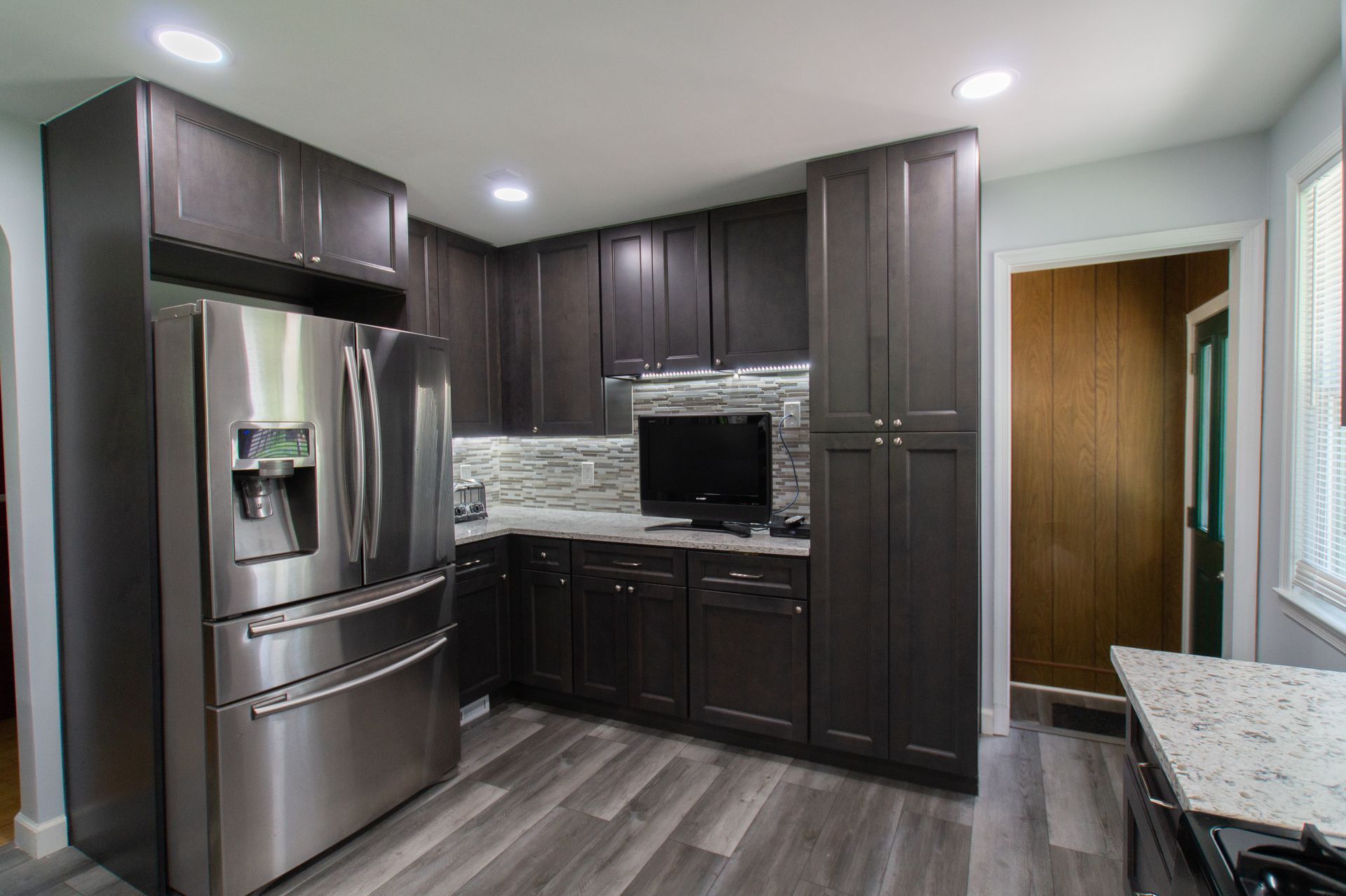 Modern kitchen with dark brown cabinets, stainless steel refrigerator, and wood-look flooring.