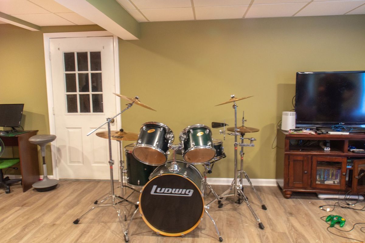 Drum set in a room with a TV and a door, set against a green wall and wooden floor.