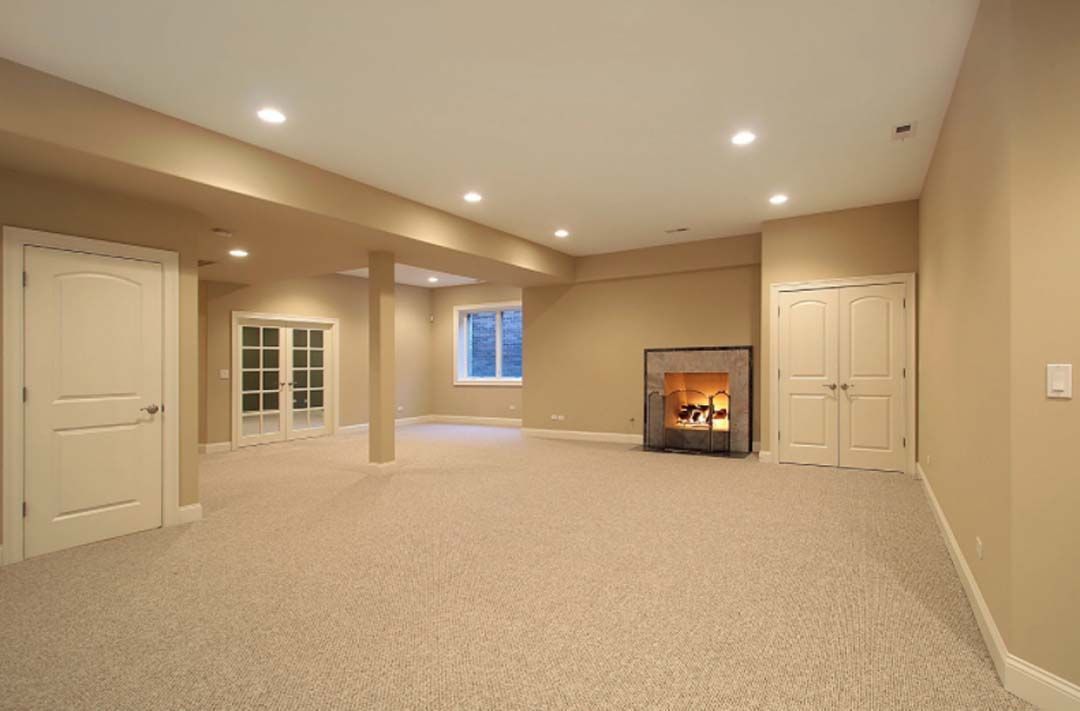 Empty beige-carpeted basement room with fireplace, doors, and recessed lighting.