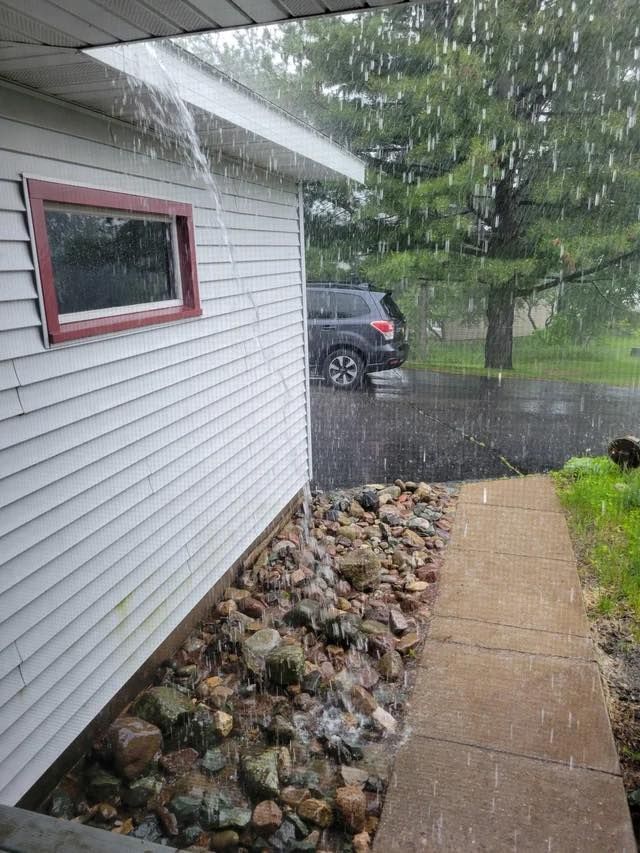 Rain pouring off the roof of a white house with a red-trimmed window, onto a rock bed and sidewalk. A car sits in the street.