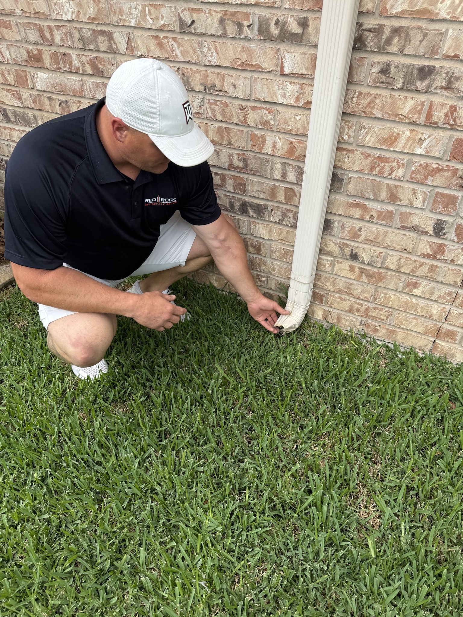 Person in shorts and cap examines a gutter downspout next to a brick wall on grass.