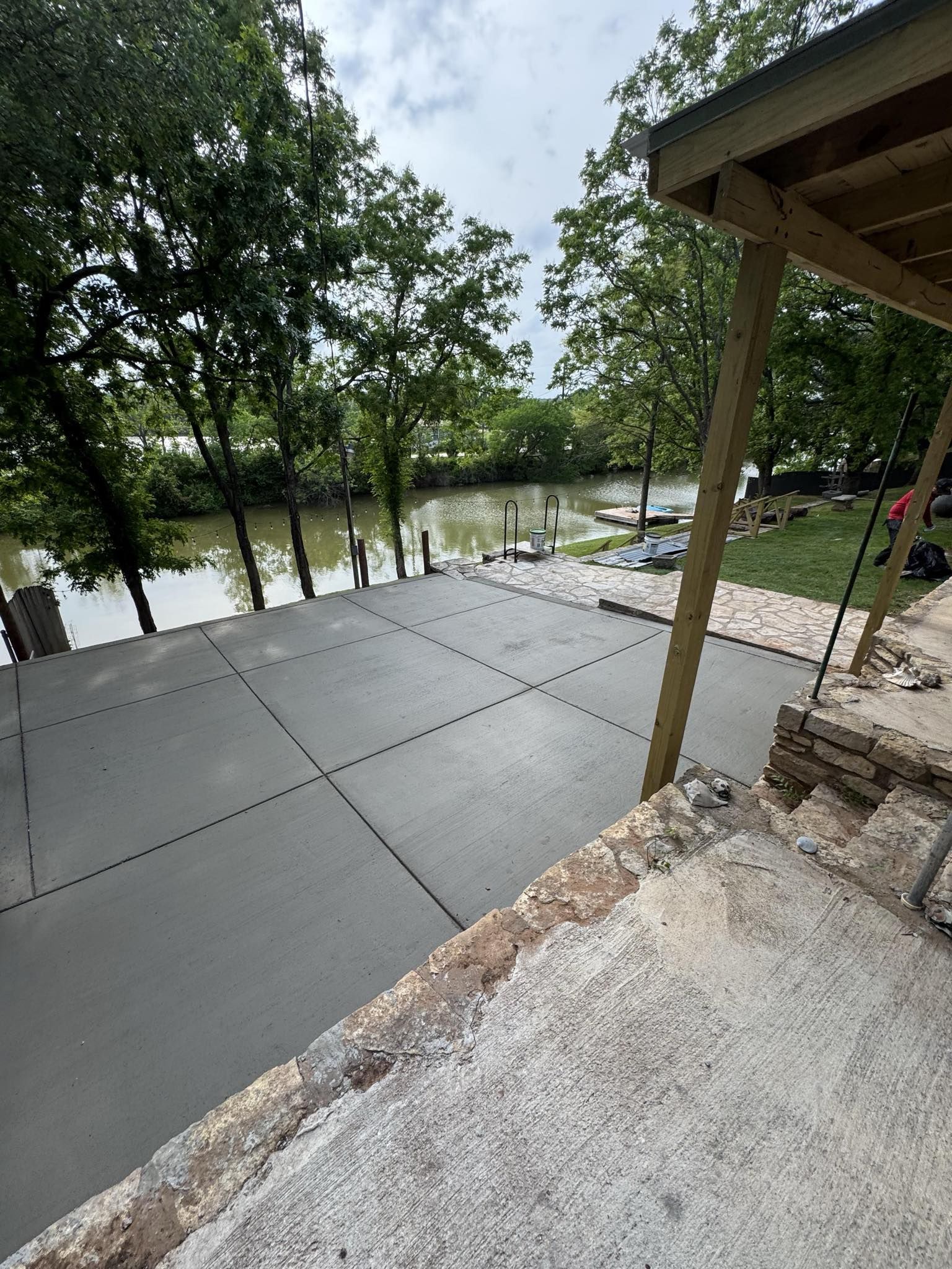 New concrete patio next to a river. Deck on right, trees in the background, overcast sky.