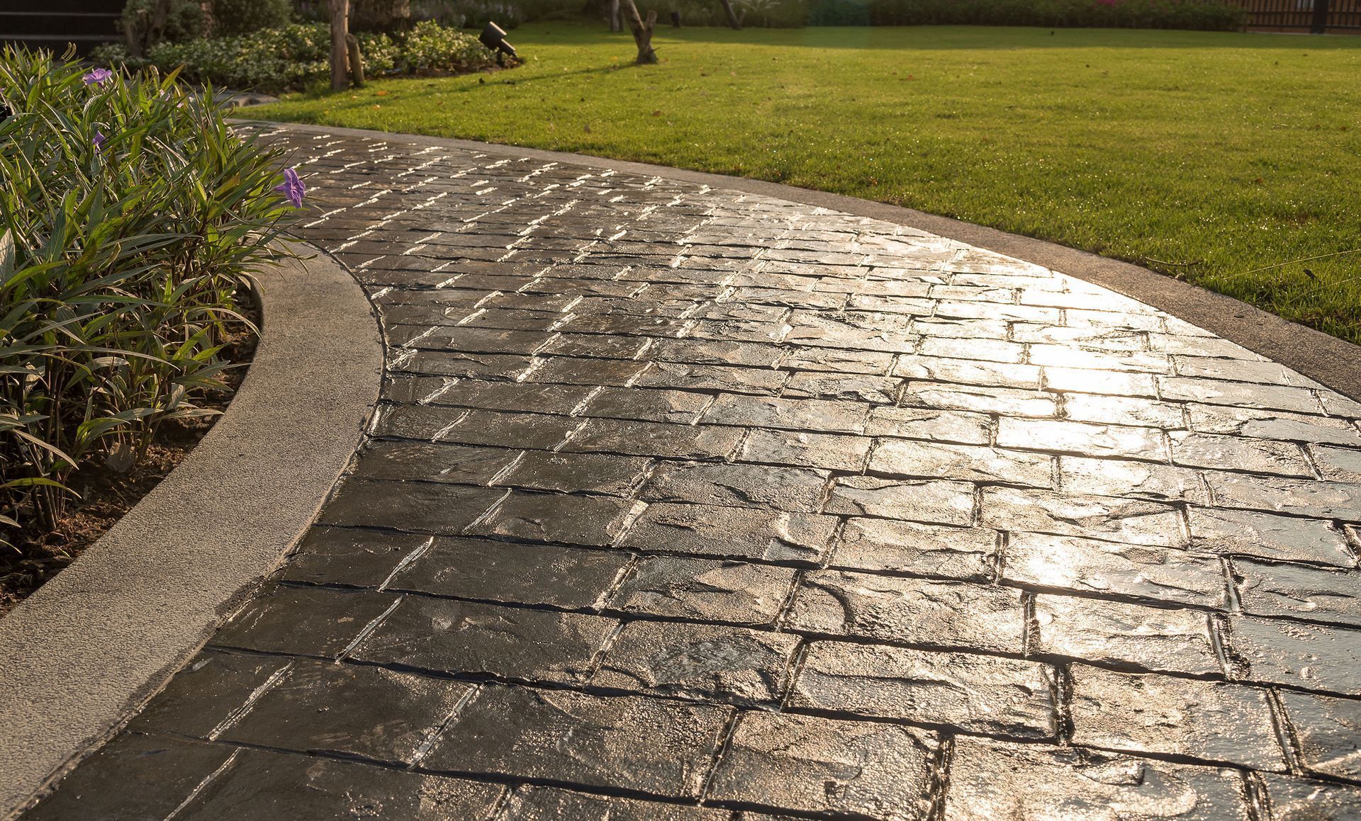 Stone-patterned walkway curves through a green lawn, bordered by plants and a concrete edge.