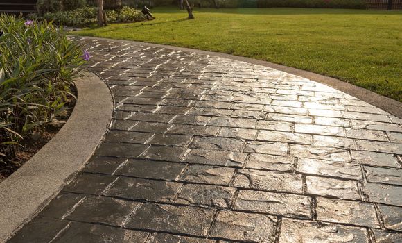 Curved, brick-patterned concrete walkway borders green grass and foliage; sunny outdoor setting.