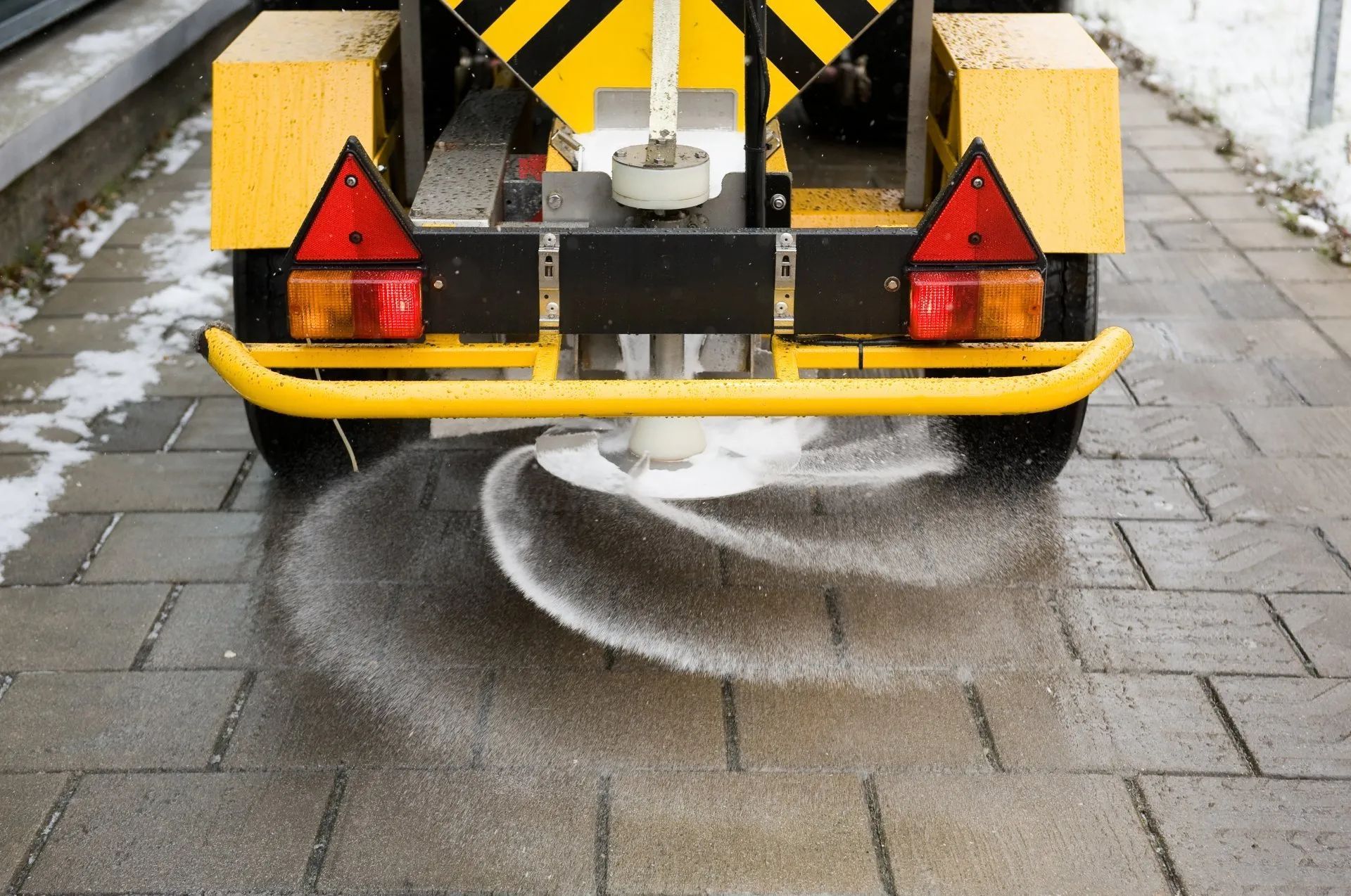A yellow and black vehicle is spreading salt on a sidewalk.