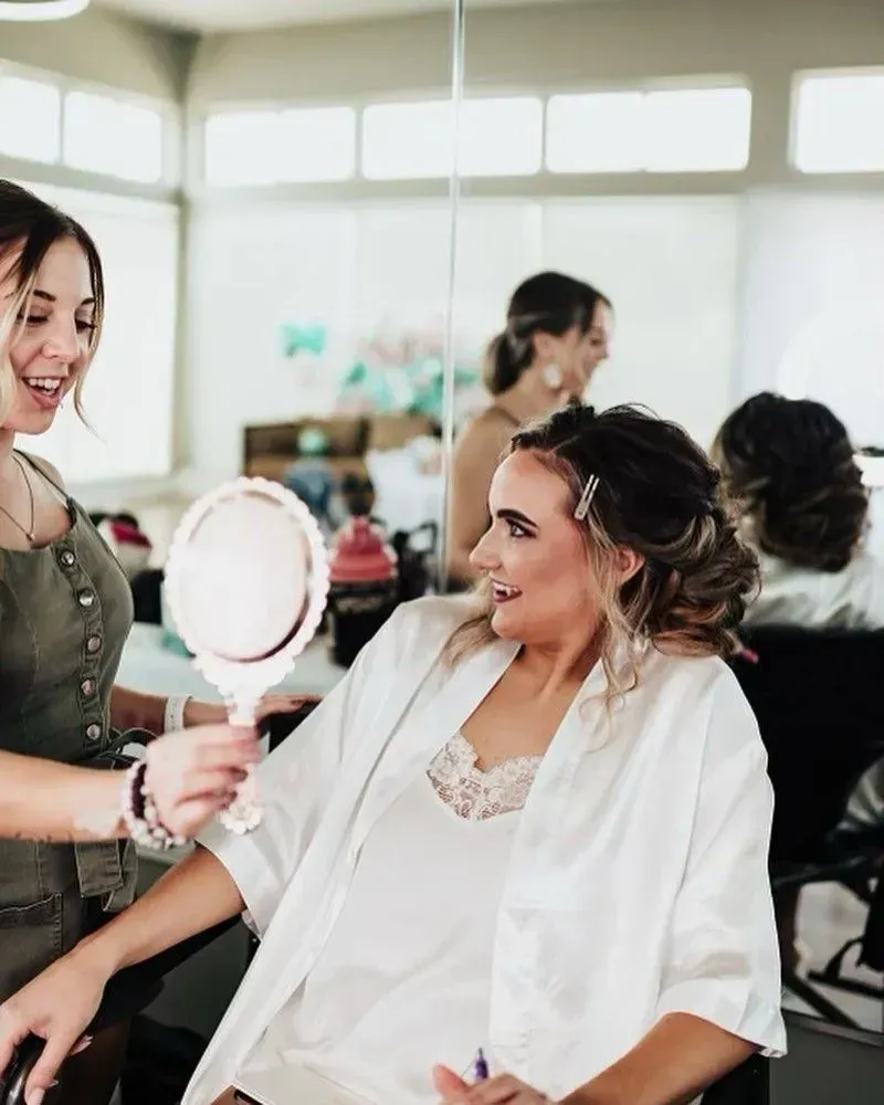 Woman in white robe smiling in a salon chair while another woman holds a hand mirror nearby