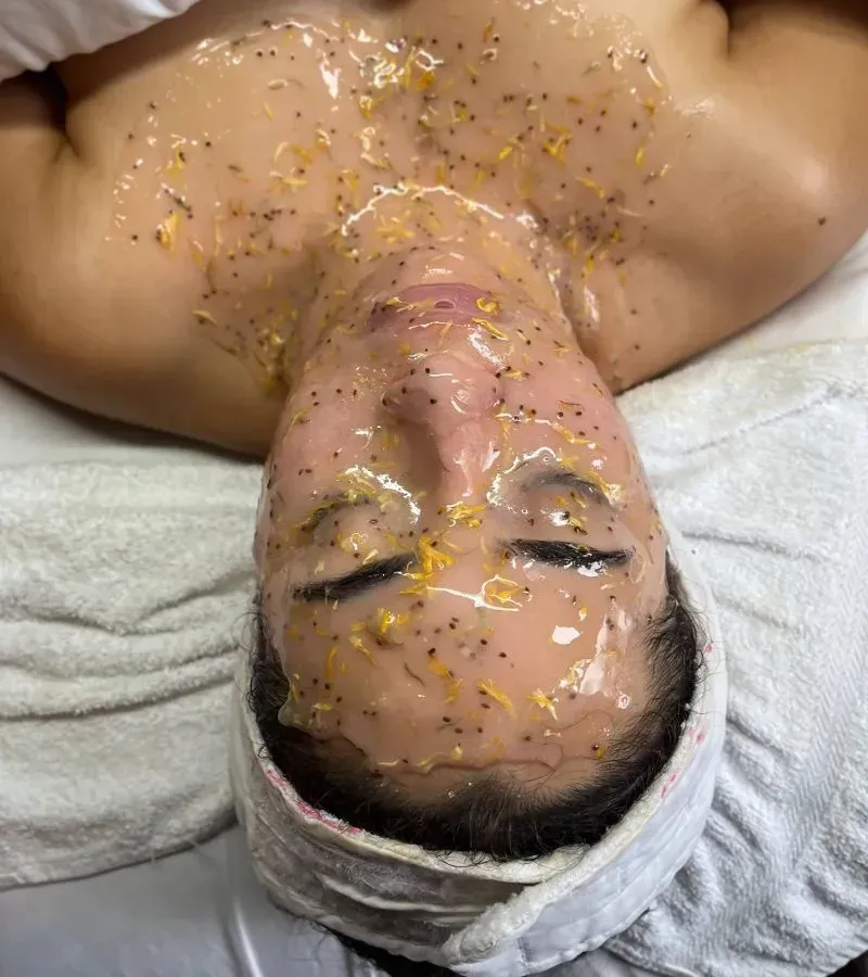 Person lying on a treatment bed with a facial mask and head wrapped in a towel, eyes closed