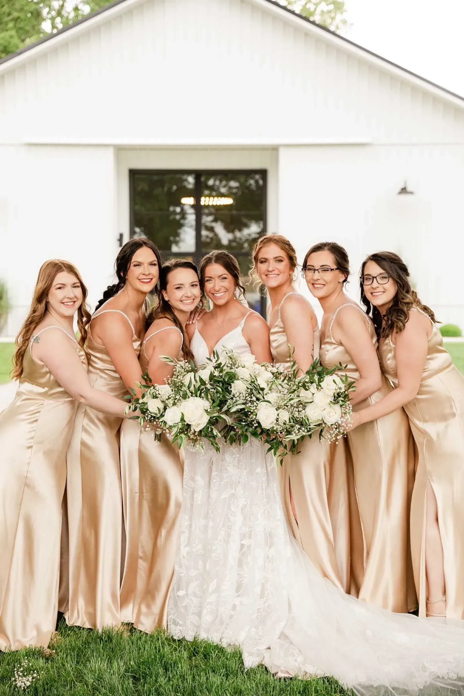 Bride and bridesmaids in champagne dresses holding a white floral bouquet in front of a white barn