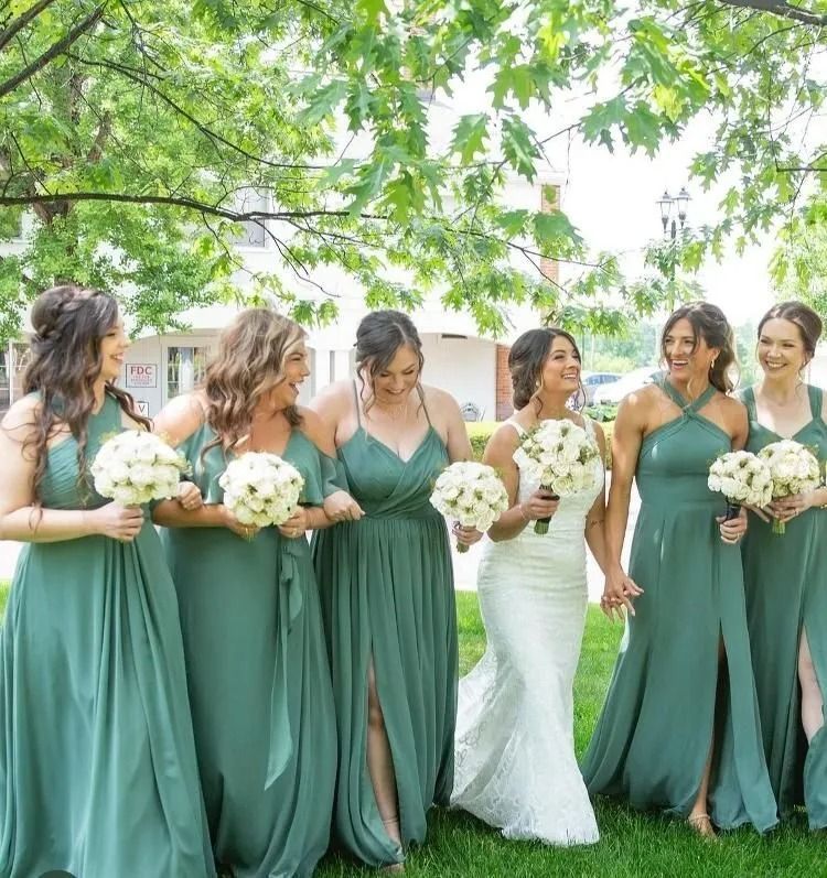 Bridesmaids in green dresses and bride in white holding bouquets under leafy trees outdoors