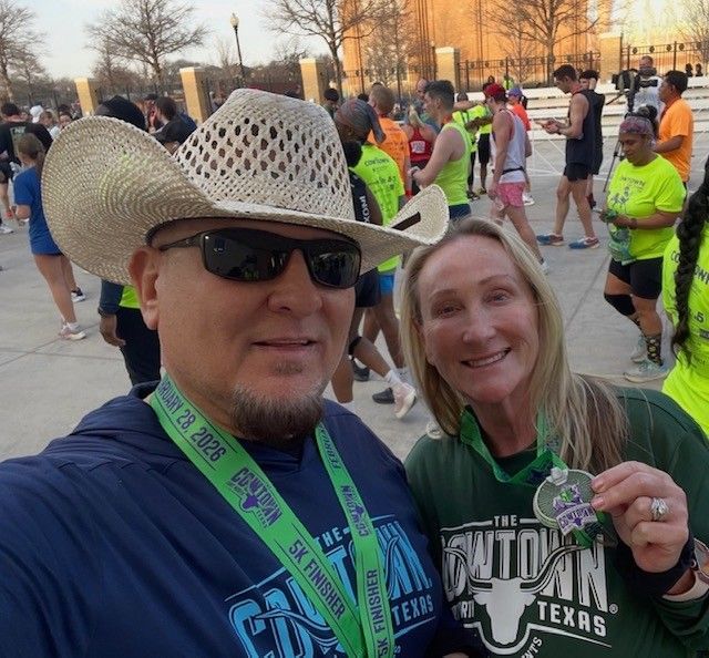 Selfie of two runners at a race event, one wearing a cowboy hat and medals, with others in the background