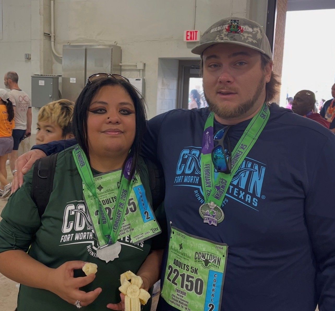 Two people in a gym smiling with medals and event bibs, standing close together.