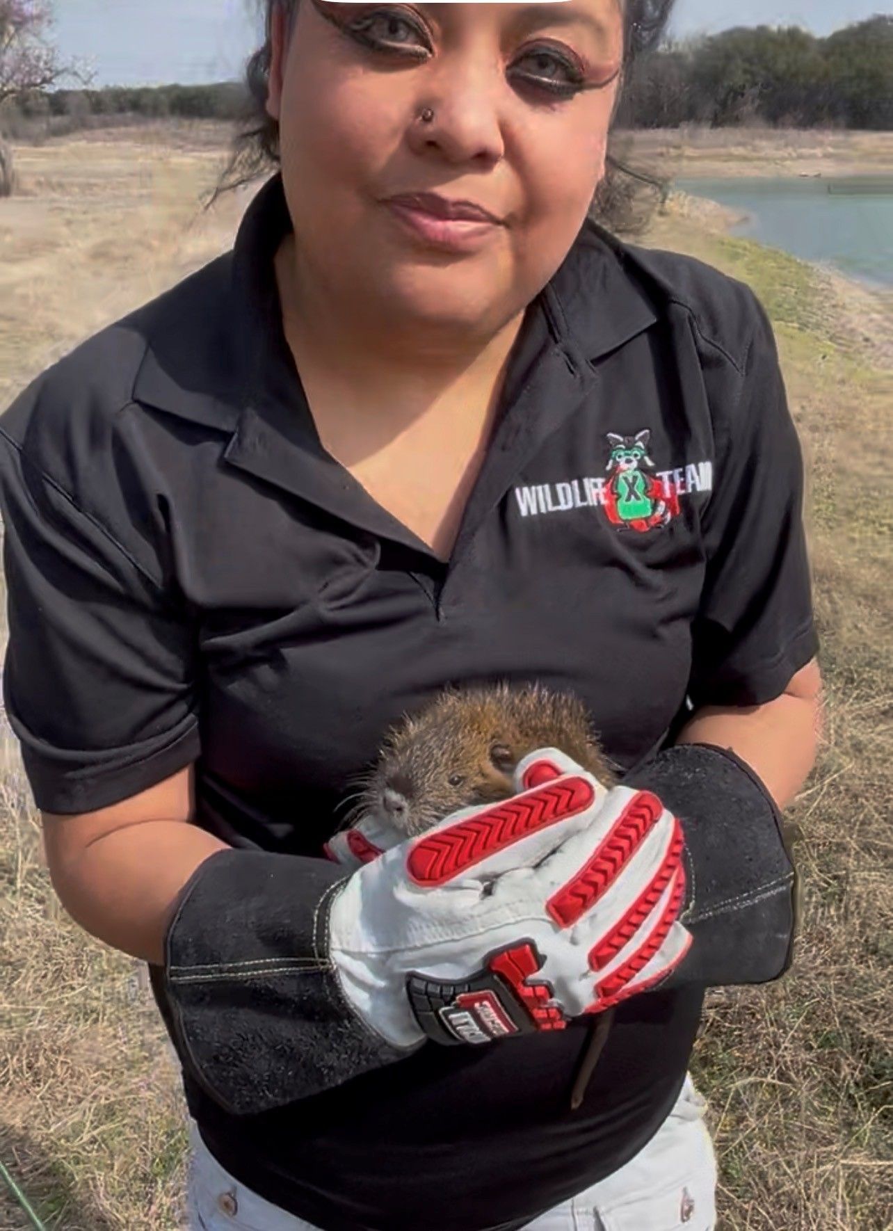 Person in a black polo shirt holding a small animal near a riverbank.