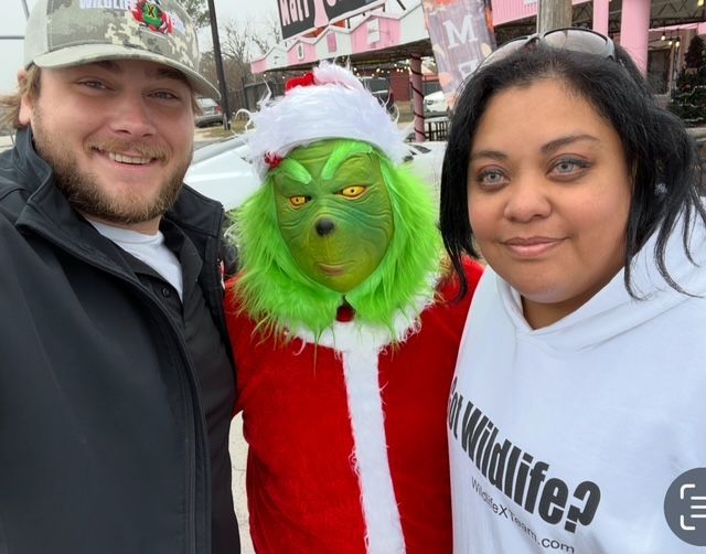 Three people posing at a holiday event, including one in a Grinch costume and red Santa outfit.