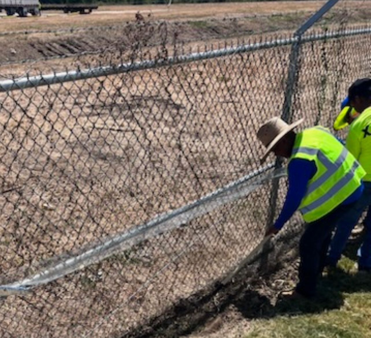 Two workers in neon vests repairing a chain-link fence beside a dry field