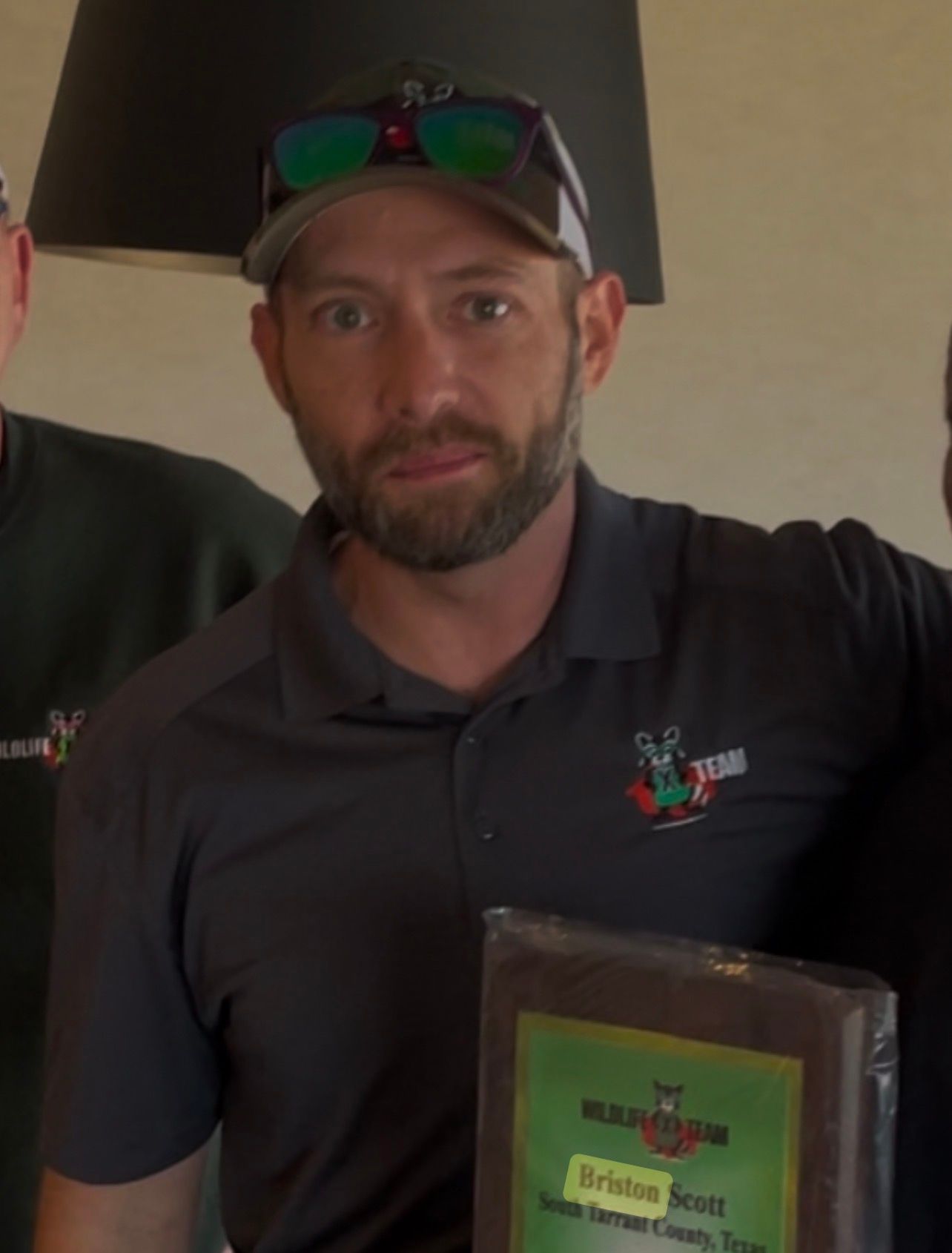 Man in dark polo and cap holds a framed plaque, standing indoors and facing the camera.