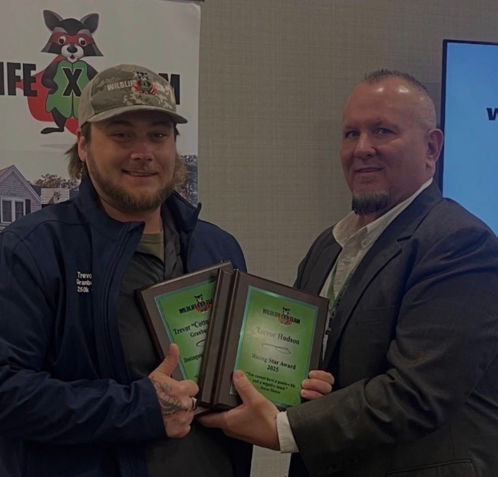 Two men in suits and cap hold framed awards at an indoor event booth.