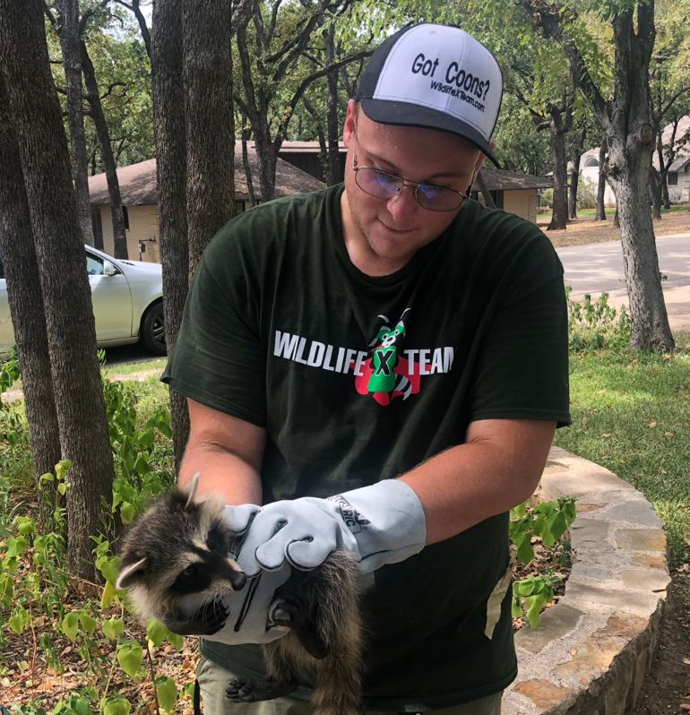 Man in cap and glasses holding a raccoon outdoors on a wooded path