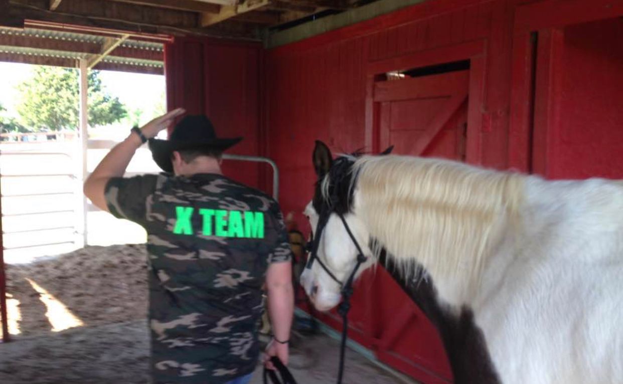 Person in black cowboy hat stands by a white horse inside a red stable.