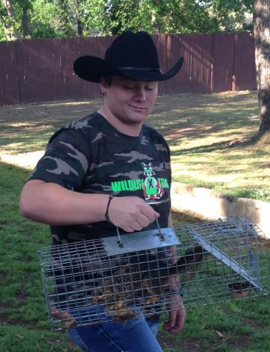 Person in a black cowboy hat holding a wire animal trap outdoors in a grassy yard.